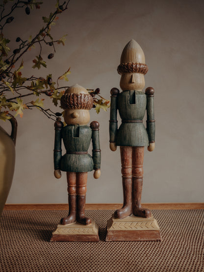 Two crafted wooden acorn nutcracker figurines, one small and one large, standing on textured bases against a neutral beige background with decorative autumn branches on the left.