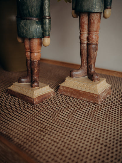 Close-up of two carved wooden soldier figurines standing on a woven brown mat, showing their legs, boots, and bases in warm natural tones.