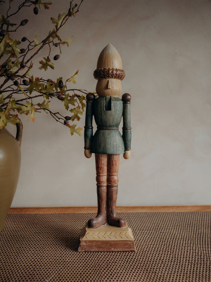 A crafted wooden nutcracker figurine with a painted teal-blue coat, brown legs, and an acorn-shaped head, standing on a decorative base against a neutral beige wall.
