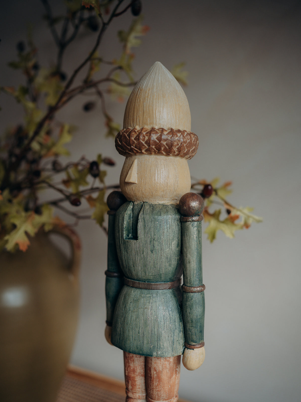 A crafted wooden nutcracker figurine shown from the back, featuring a light natural wood cone-shaped head, a braided brown band, a painted teal-green tunic, and natural wood legs, set against a soft gray background with blurred autumn leaves in a vase.