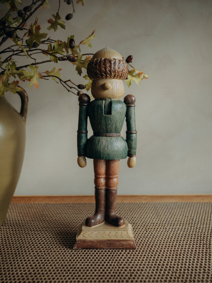 A crafted wooden nutcracker figurine with a green tunic, brown boots, and an acorn hat, standing on a textured woven mat with a vase and autumn leaves in the background.