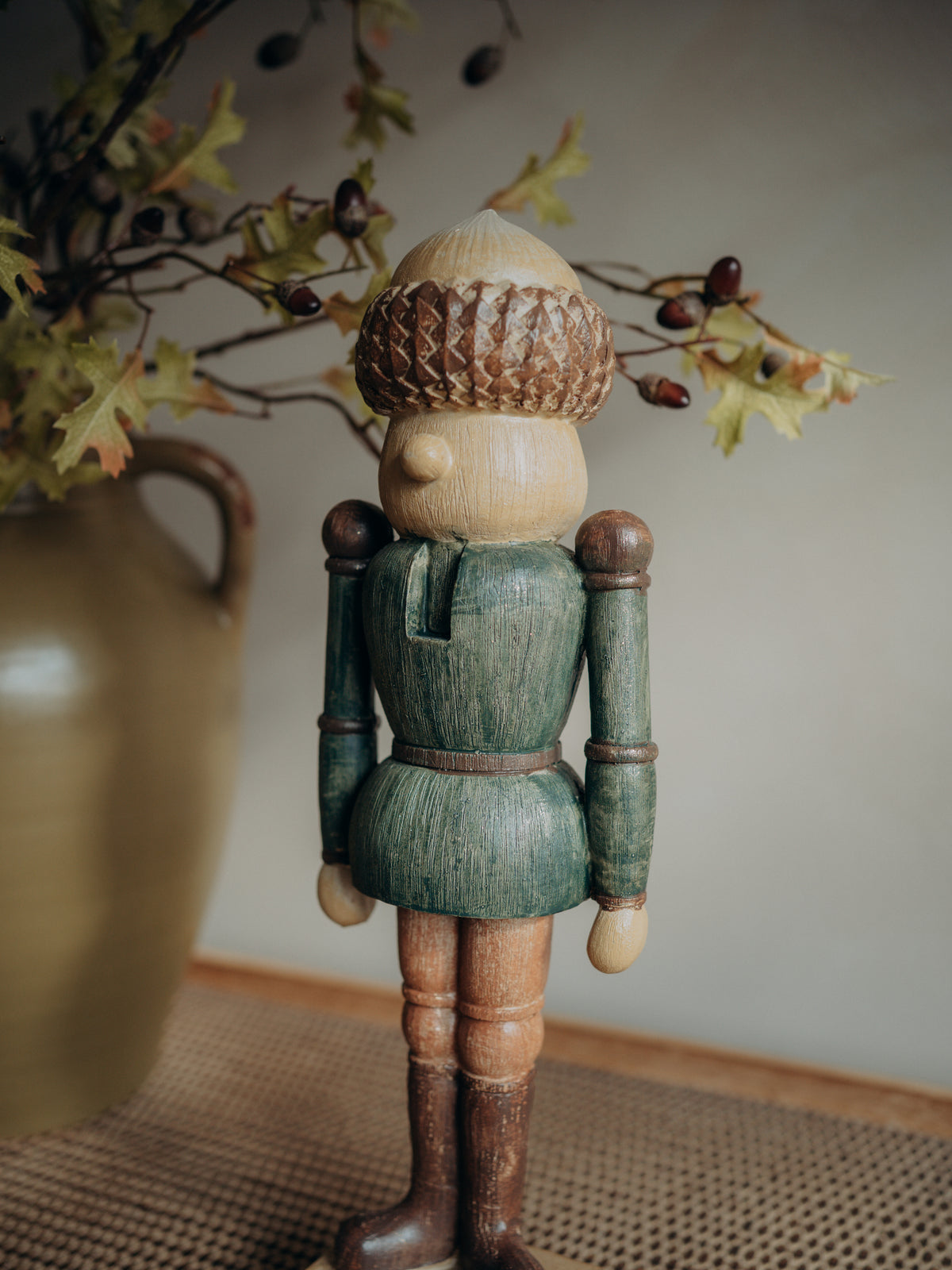 A crafted wooden nutcracker figurine with a green painted tunic, brown wooden legs and boots, and a textured acorn hat, standing on a woven mat with a blurred vase of branches in the background.