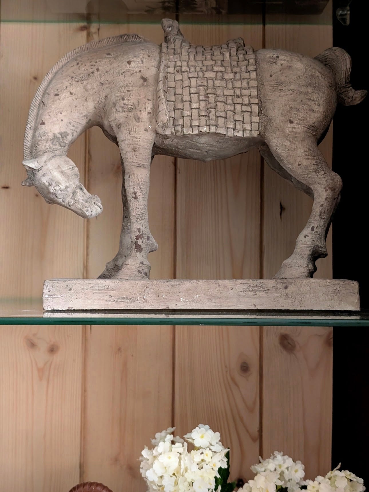 A rustic stone horse sculpture with a woven saddle, displayed on a glass shelf against a light wood paneled background.