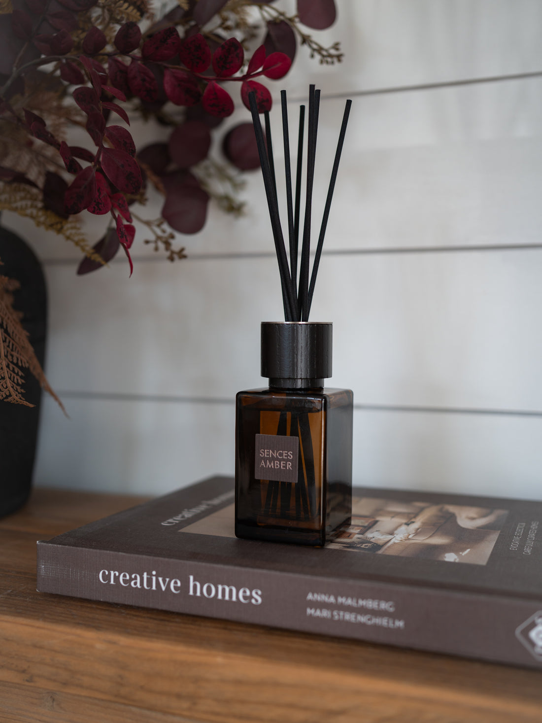 A sences amber reed diffuser bottle with black reeds sits on top of a book on a wooden surface, with dried flowers in the background against a white shiplap wall.