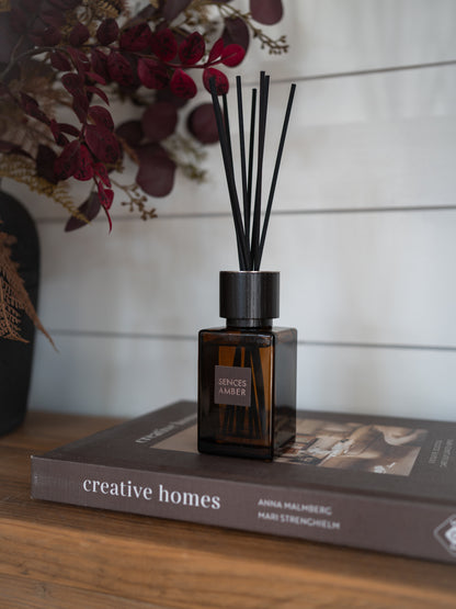 A sences amber reed diffuser bottle with black reeds sits on top of a book on a wooden surface, with dried flowers in the background against a white shiplap wall.