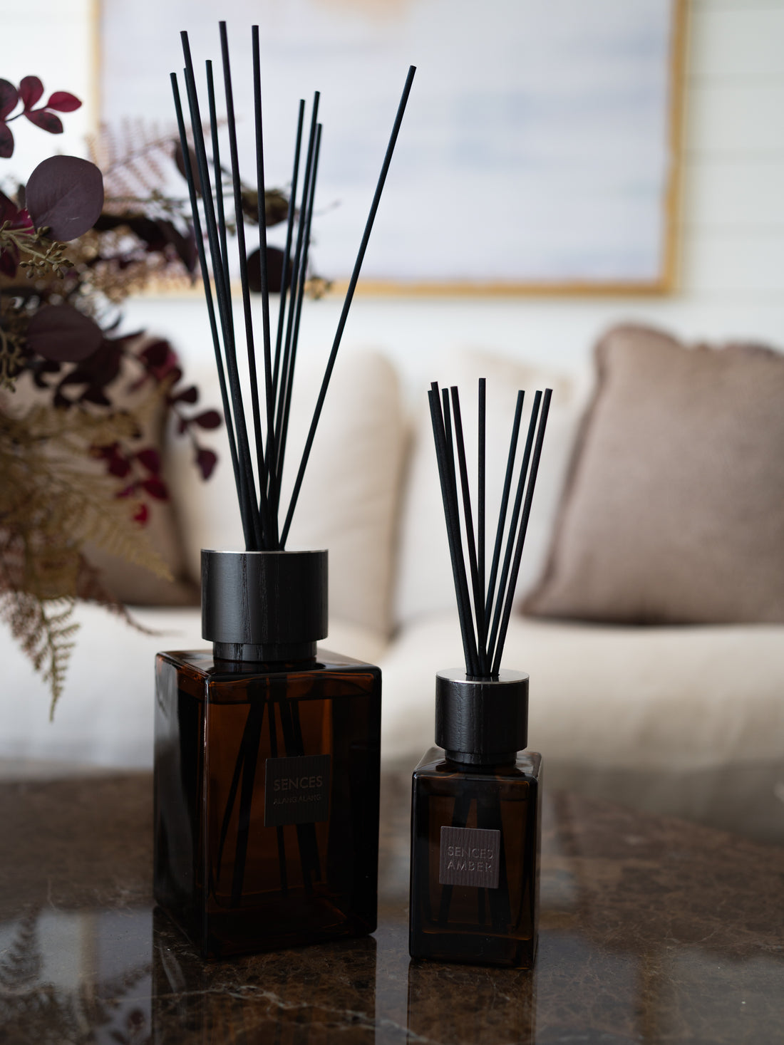Two sences reed diffusers in dark amber glass bottles with black caps and reeds, displayed on a polished marble table in a softly blurred living room setting.