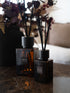 Two amber glass reed diffusers with black caps and black reeds, labeled &