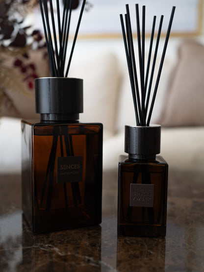 Two amber glass reed diffusers with black caps and black reeds, standing on a dark marble surface in a softly blurred indoor setting.
