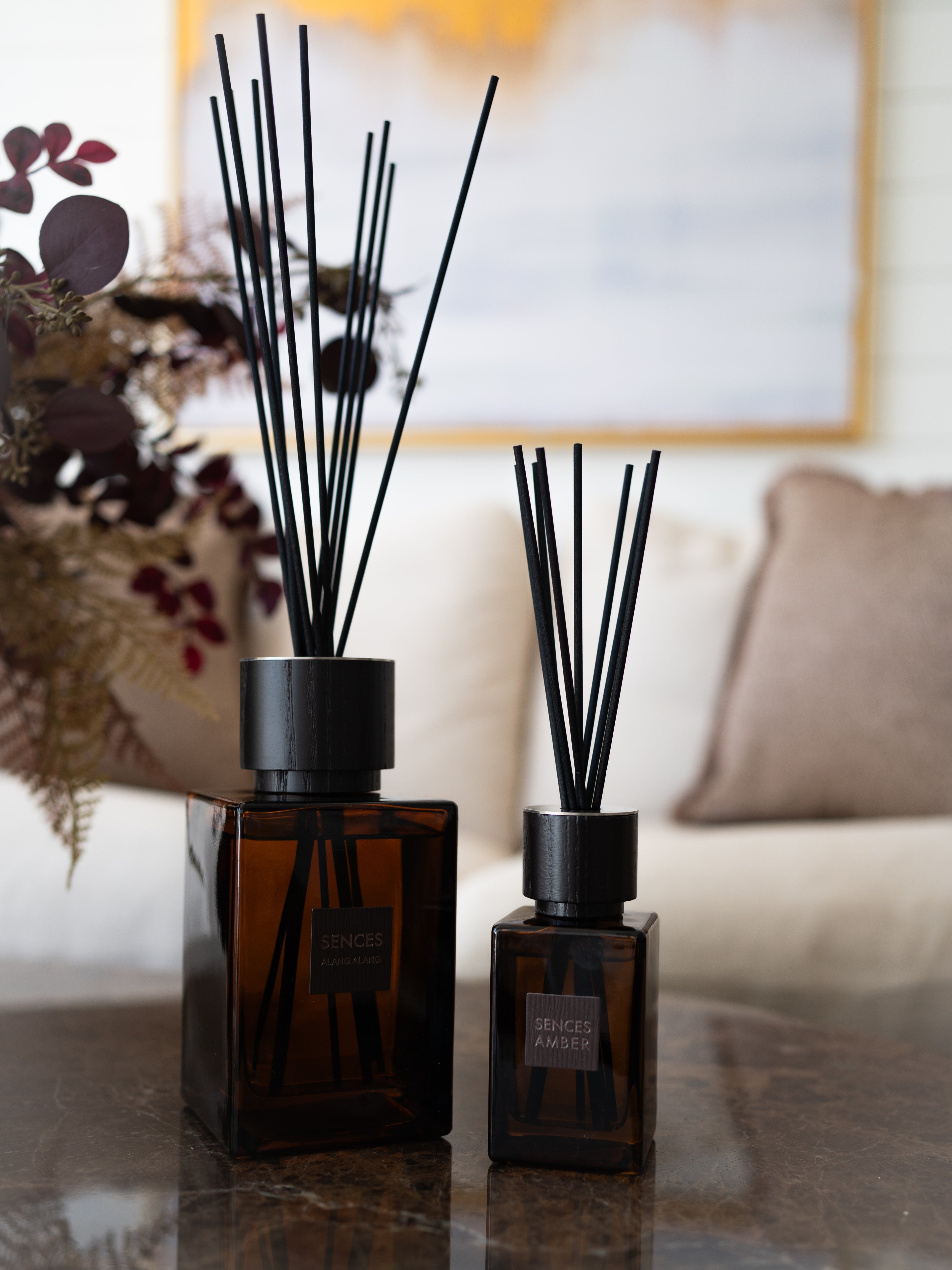 Two amber glass reed diffusers with black caps and reeds, placed on a polished stone table in a softly blurred living room setting.