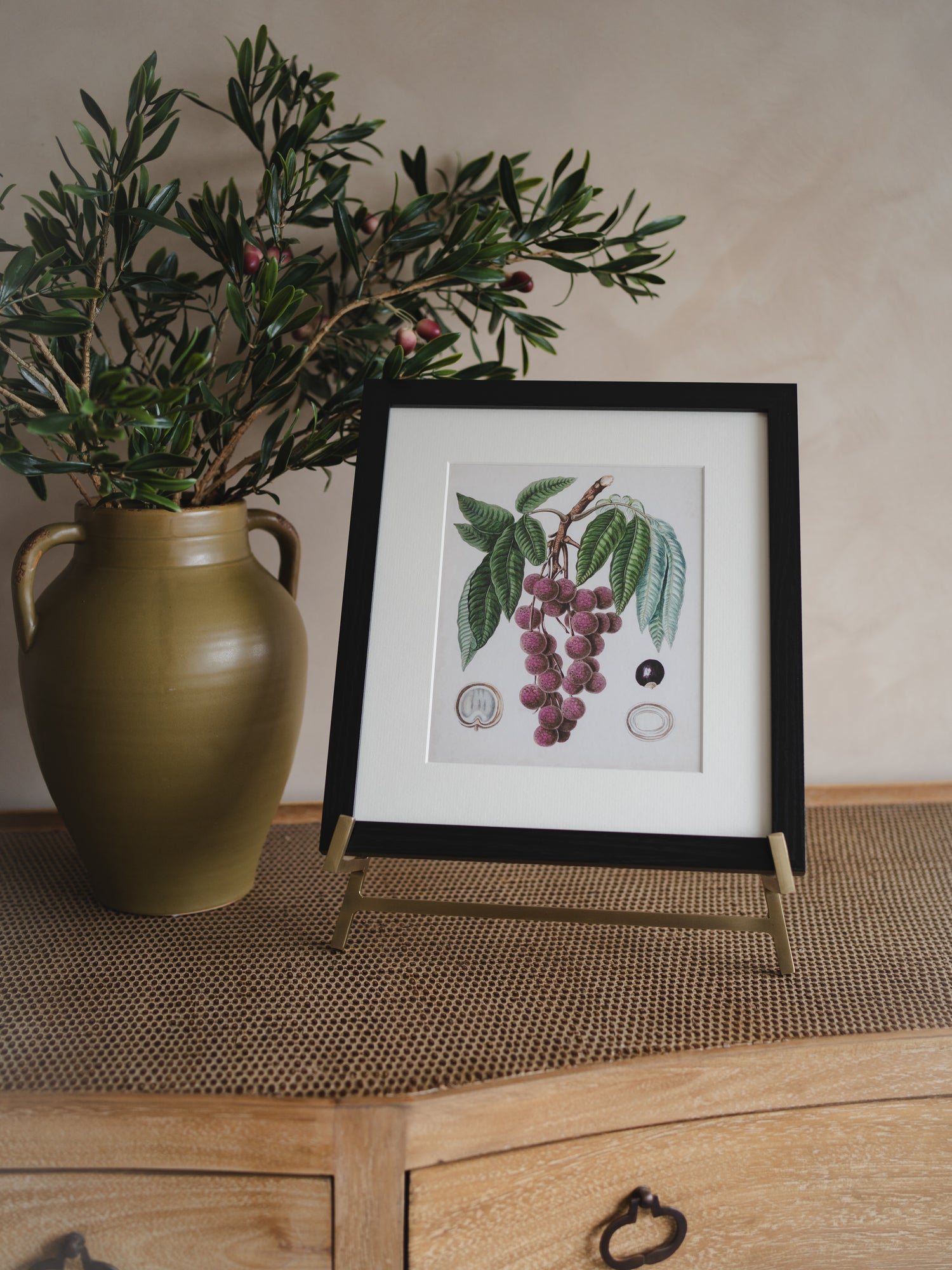 Framed botanical print on antique brass easel to a green vase with plants on a wooden surface.