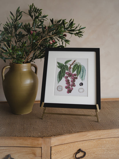 Framed botanical print on antique brass easel to a green vase with plants on a wooden surface.