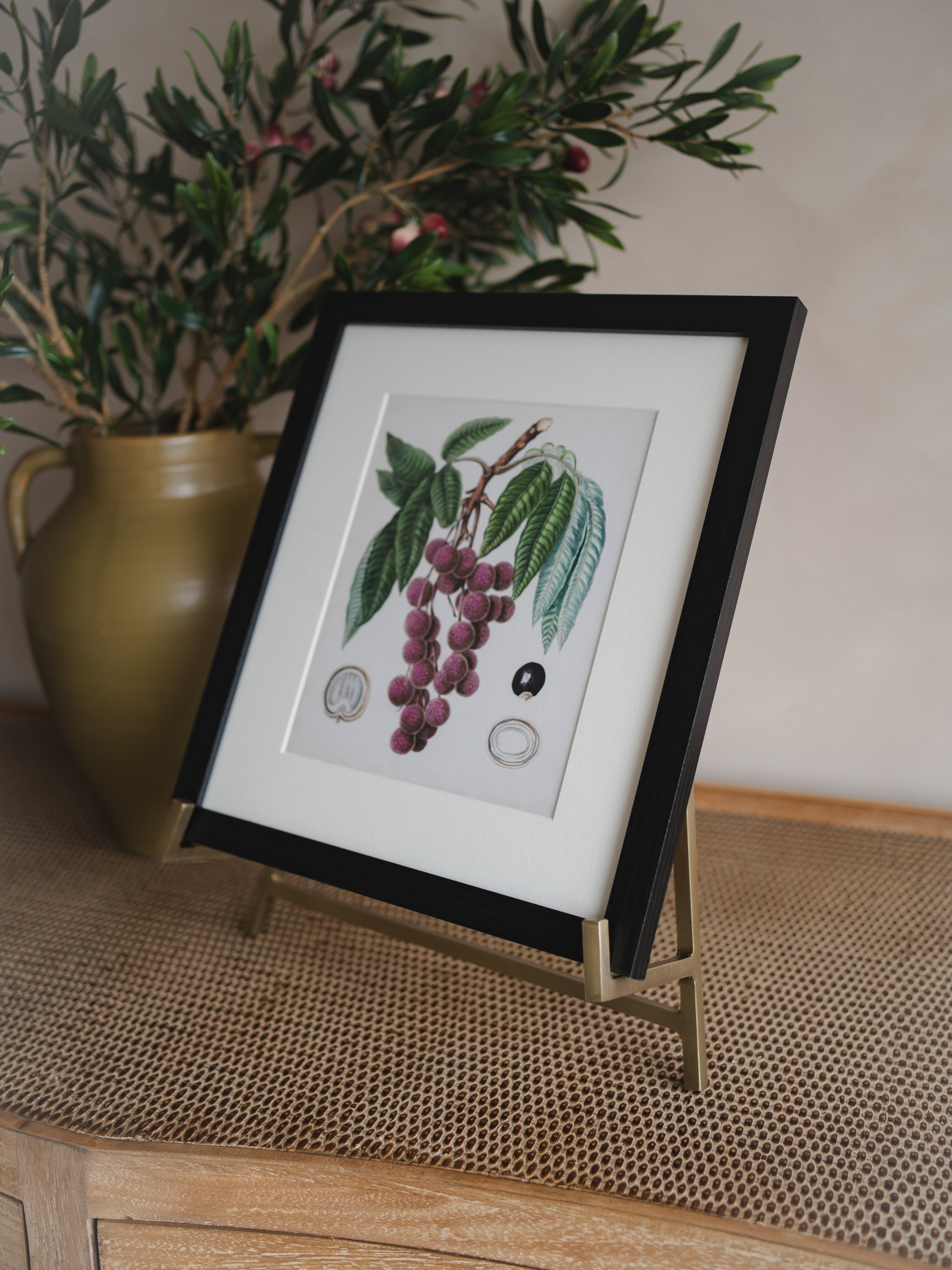 Framed botanical illustration on an antique brass easel on a table with a vase of flowers in the background