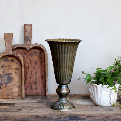 A vintage-style fluted pedestal vase in an antique bronze finish, displayed on a rustic wooden table next to a white flower pot with green plants and two wooden cutting boards against a plain off-white wall.