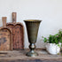 A vintage-style fluted pedestal vase in an antique bronze finish, displayed on a rustic wooden table next to a white flower pot with green plants and two wooden cutting boards against a plain off-white wall.