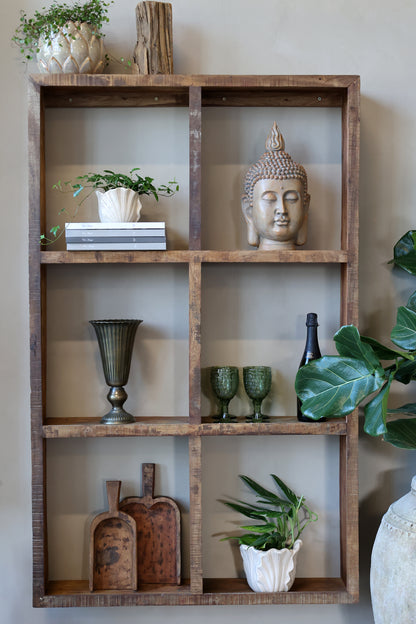 A rustic wooden floating shelf with six open cubbies, styled with home decor items including a Buddha statue, potted plants, books, and glassware, set against a neutral beige wall.