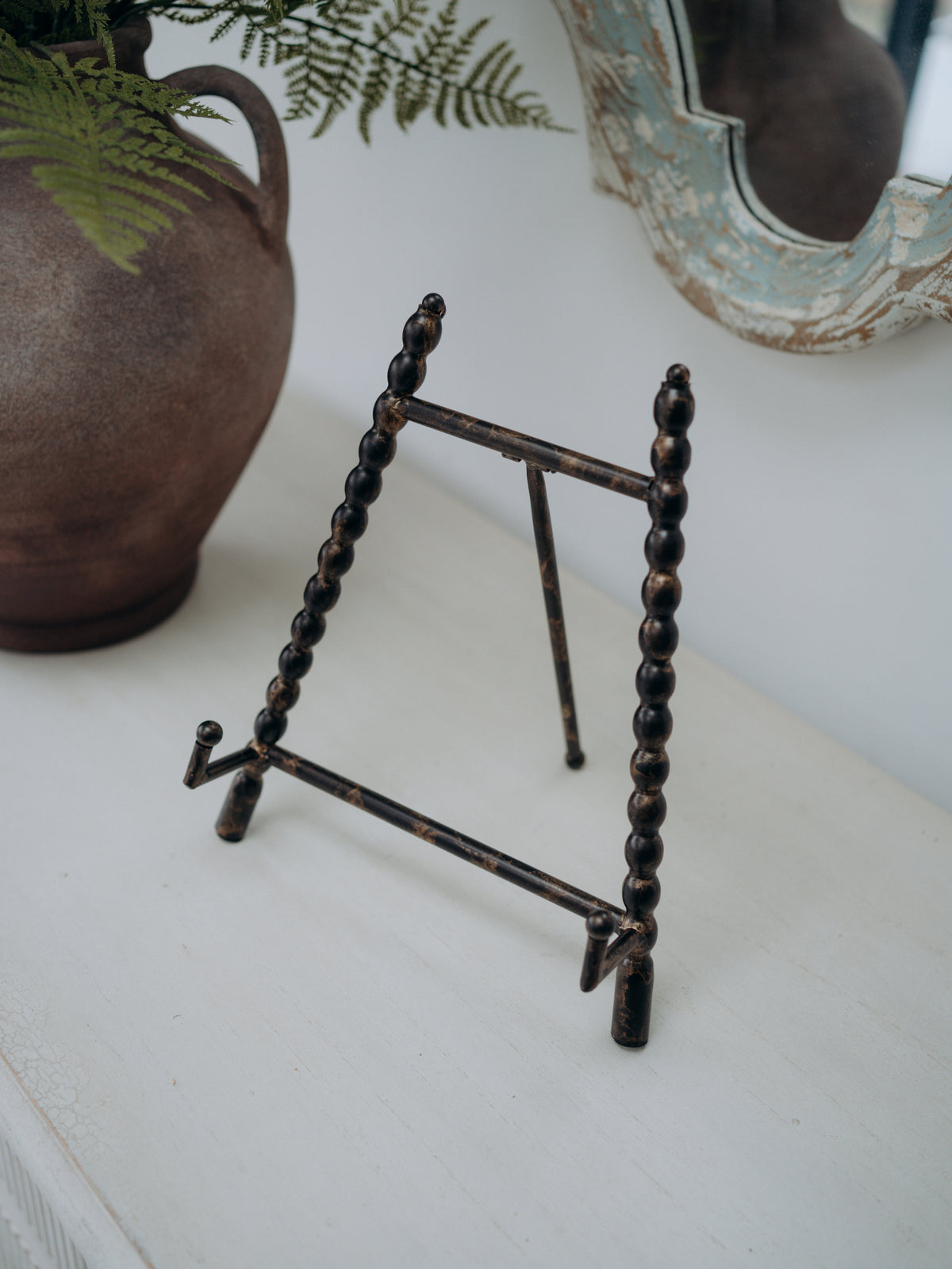 A small rustic metal display easel stand with dark brown and bronze tones, styled on a white tabletop next to a ceramic vase with green ferns.
