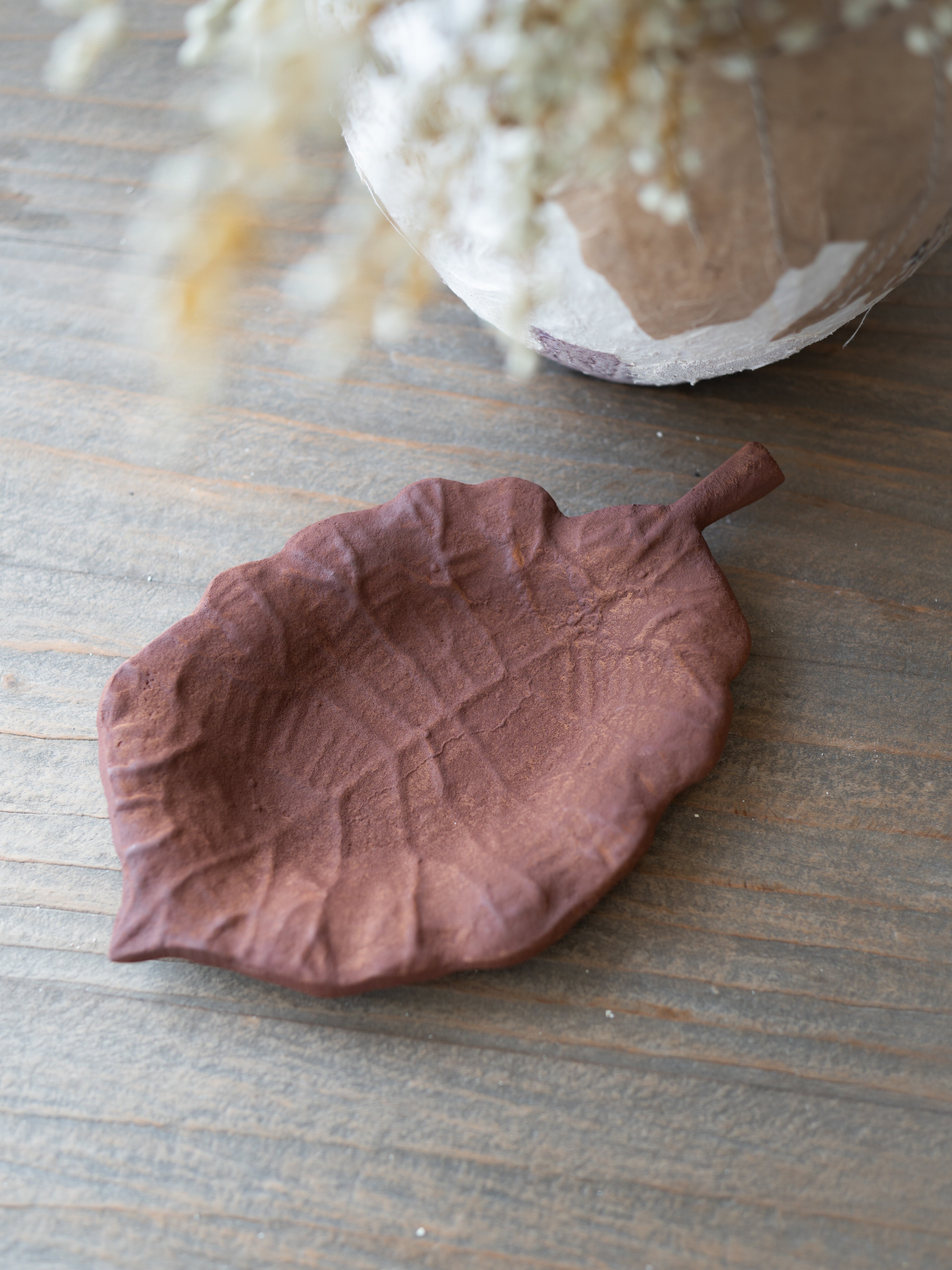 A leaf-shaped ceramic dish in a dusty terracotta brown color, styled on a rustic wooden tabletop with a blurred vase of dried flowers in the background.