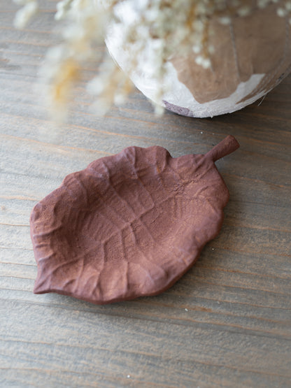 A leaf-shaped ceramic dish in a dusty terracotta brown color, styled on a rustic wooden tabletop with a blurred vase of dried flowers in the background.