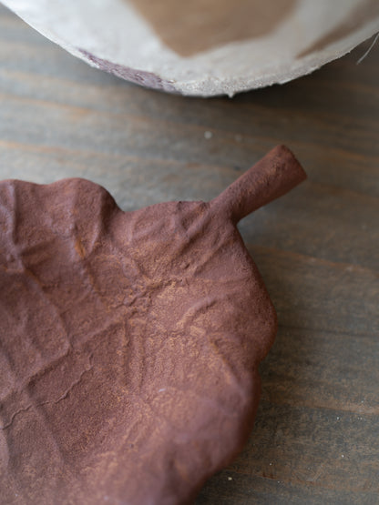 A close-up of a brown, textured leaf-shaped tray on a dark wooden surface, with part of a white bowl visible at the top edge.