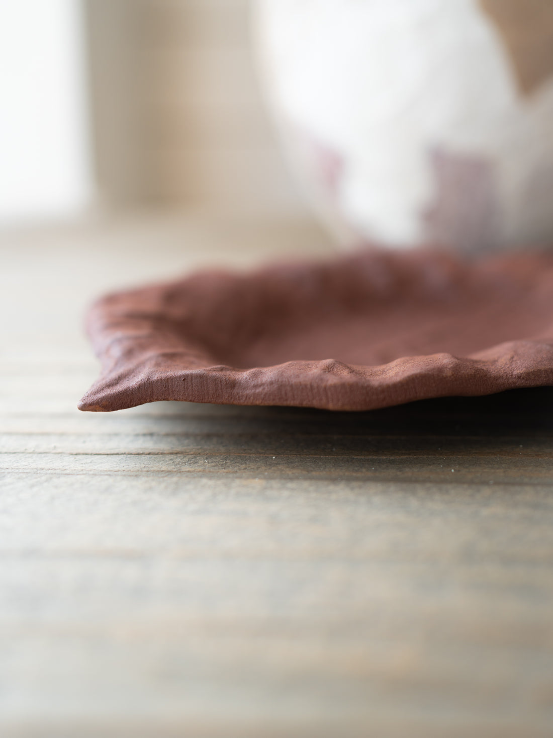 A close-up of a rustic, handcrafted terracotta-colored metal plate with a wavy rim, resting on a weathered wooden tabletop with a soft-focus background.