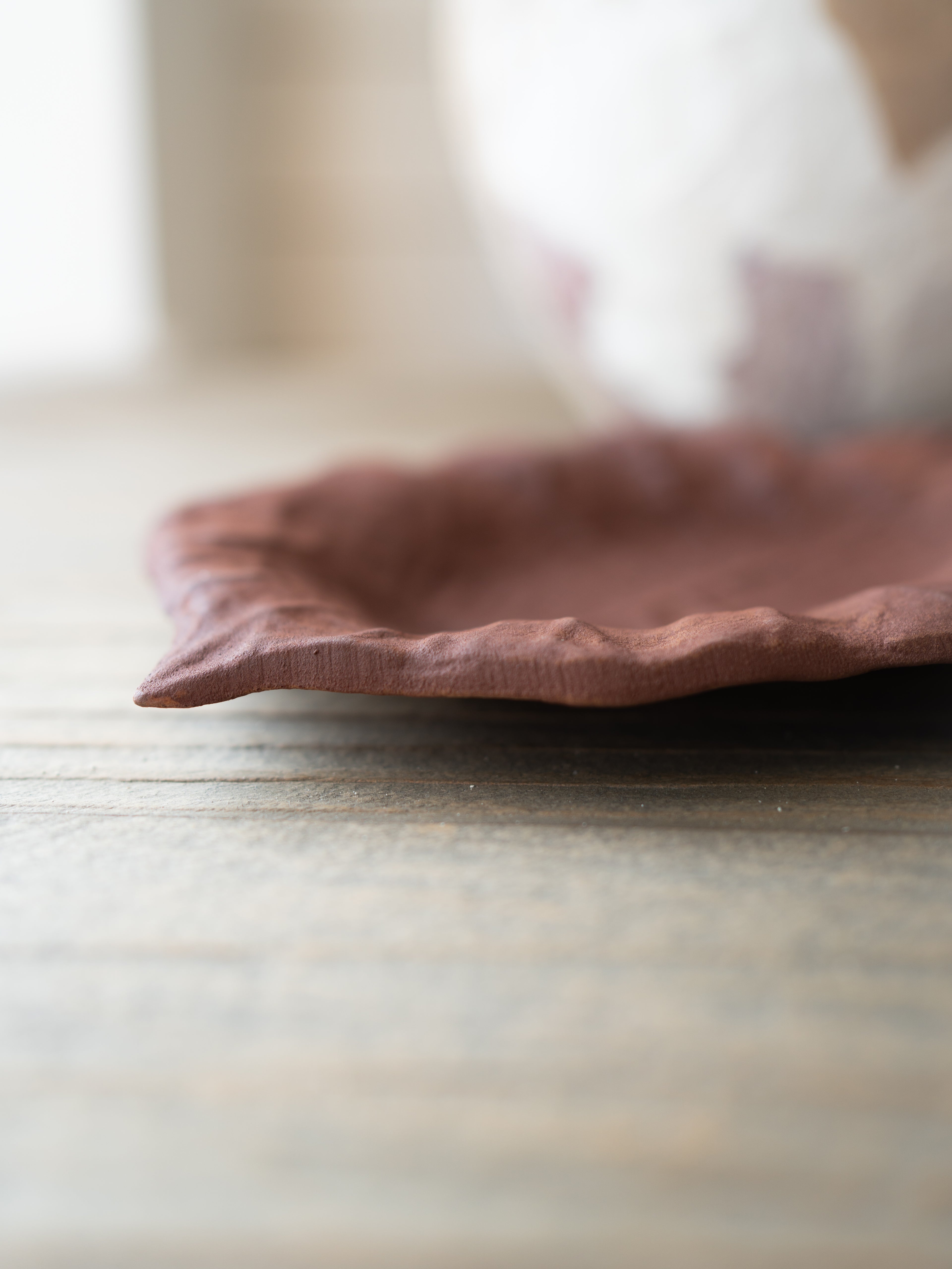 A close-up of a rustic, handcrafted terracotta-colored metal plate with a wavy rim, resting on a weathered wooden tabletop with a soft-focus background.