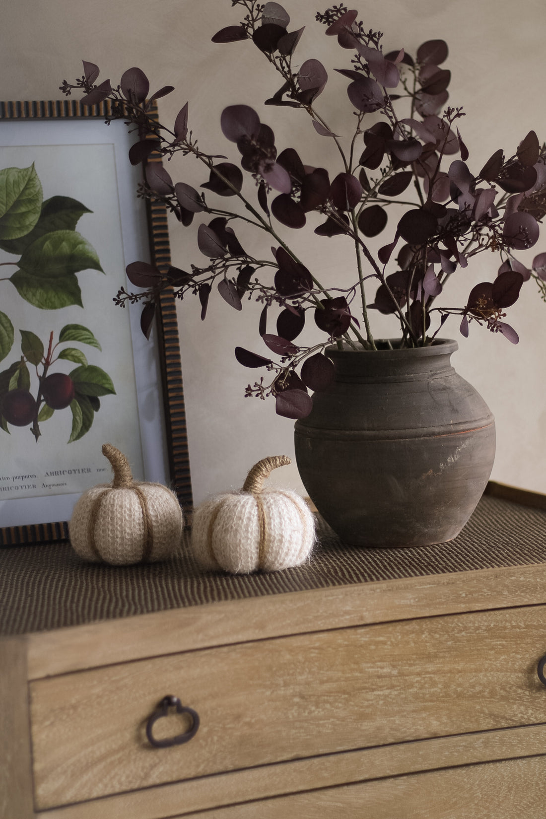 Decorative setup with a vase of purple faux eucalyptus, knitted pumpkins, and a framed botanical print on a wooden surface.