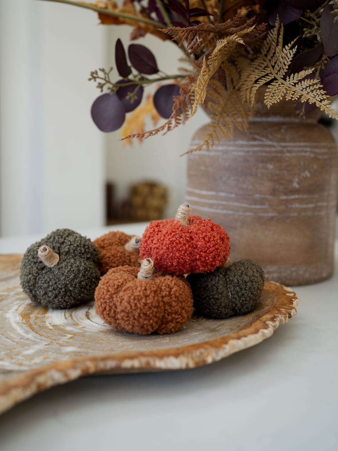 Three textured knit decorative pumpkins in rust orange, terracotta brown, and charcoal gray, arranged on a rustic wood slice tray with a blurred floral arrangement in the background.