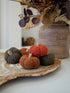 Three textured knit decorative pumpkins in rust orange, terracotta brown, and charcoal gray, arranged on a rustic wood slice tray with a blurred floral arrangement in the background.