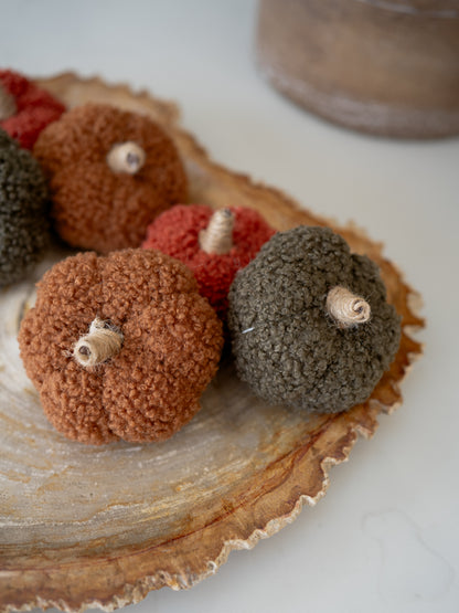 Three textured fabric pumpkins in rust orange, dark olive green, and terracotta red, arranged on a rustic wood slice platter.