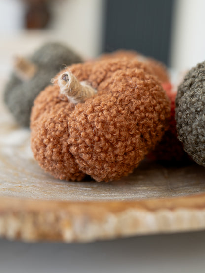 A close-up of a teddy fabric decorative pumpkin in a warm rust-brown color, placed on a wooden slice tray with other muted pumpkins in the background.