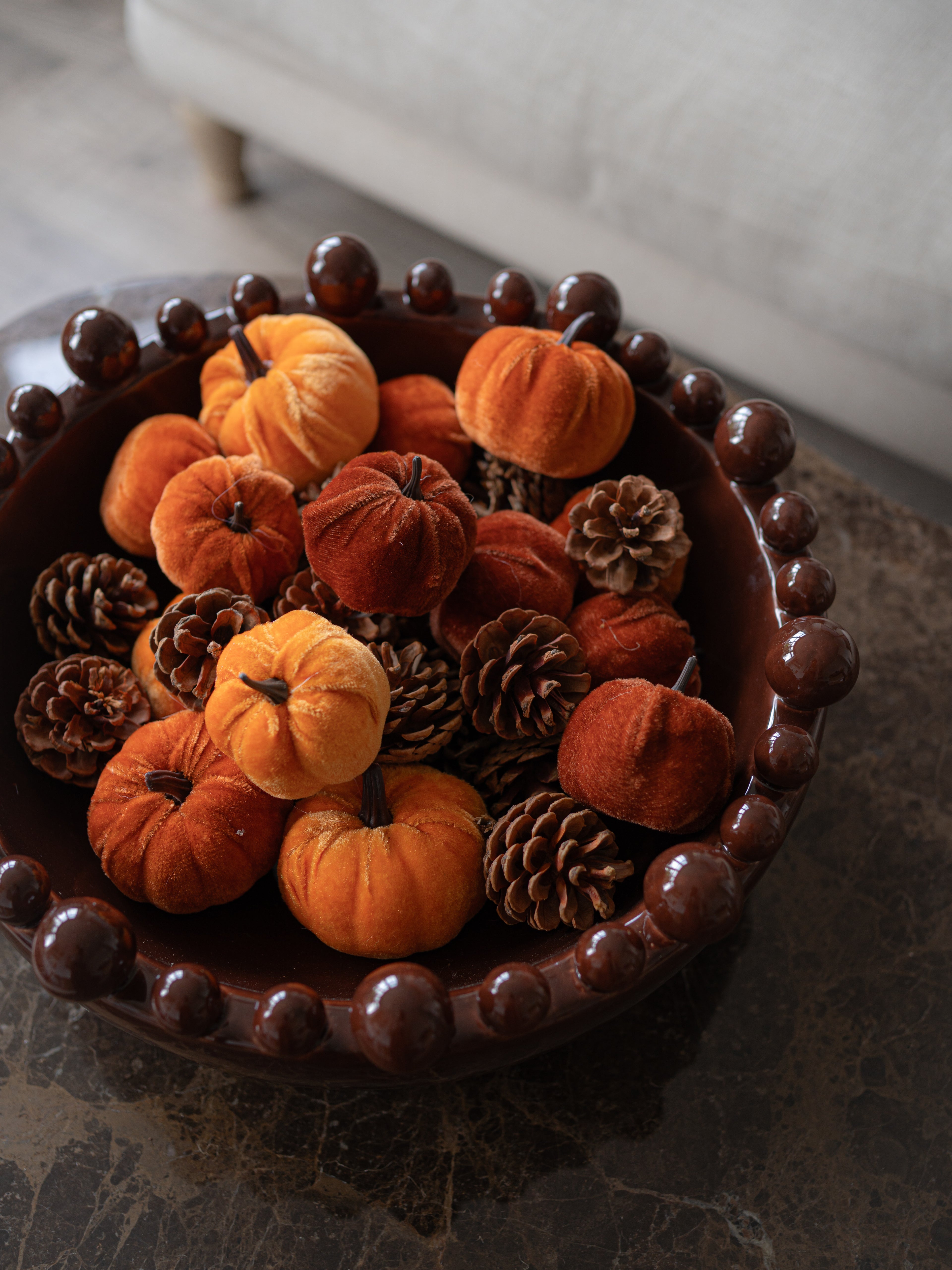 Bag of Velvet Autumn Orange Pumpkins and Rustic Pine Cones in coco brown ceramic bobble edged bowl on brown marble table