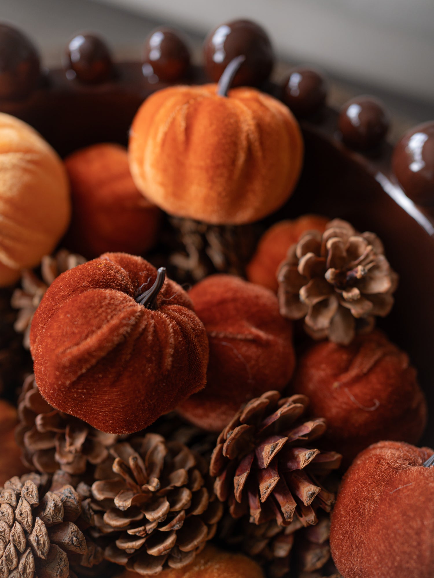 Autumn decorative arrangement featuring small velvet pumpkins in rust, orange, and terracotta tones, mixed with natural pinecones inside a dark bowl.
