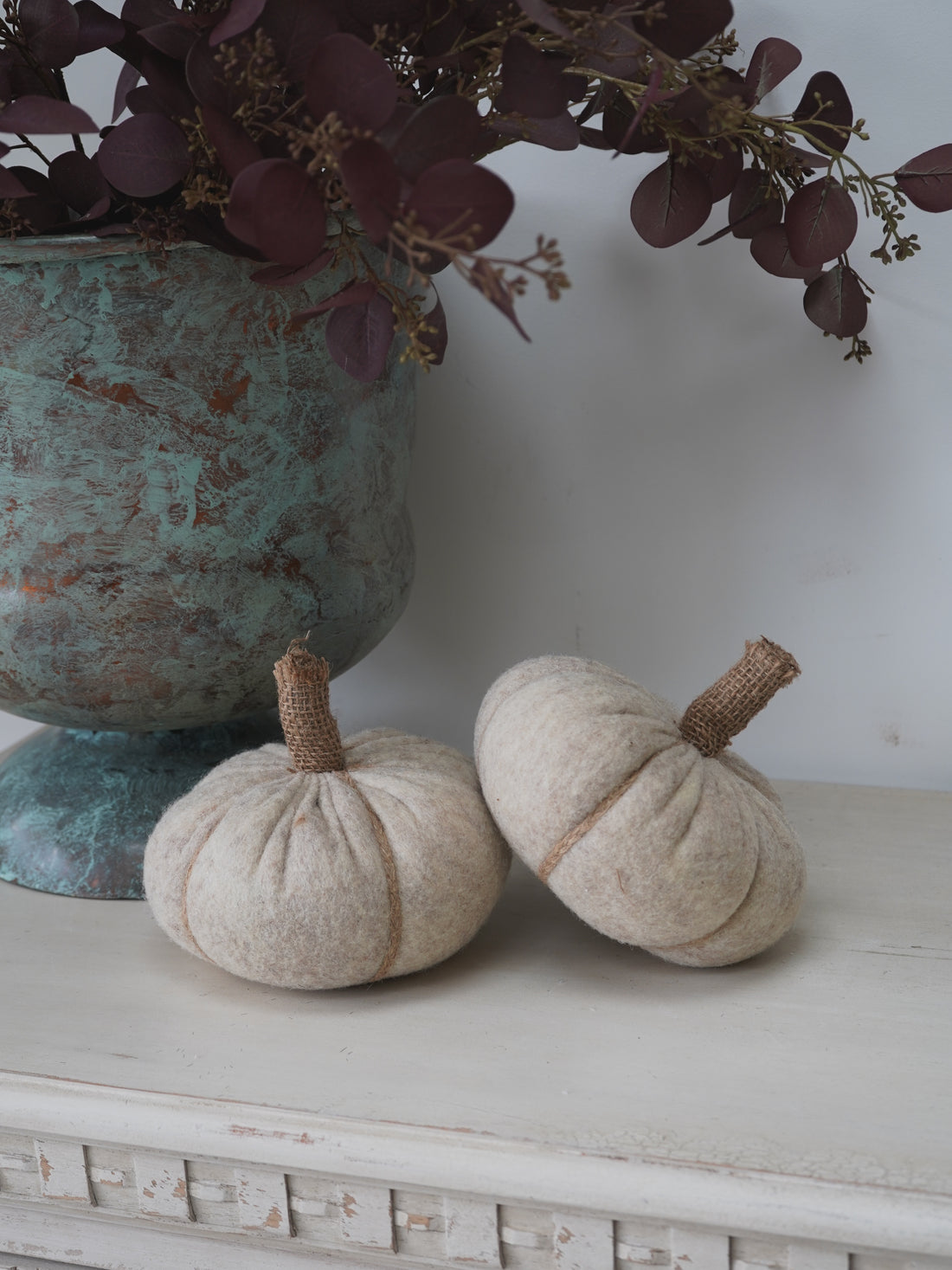 Two beige pumpkins on a white surface with a decorative urn and purple foliage in the background.