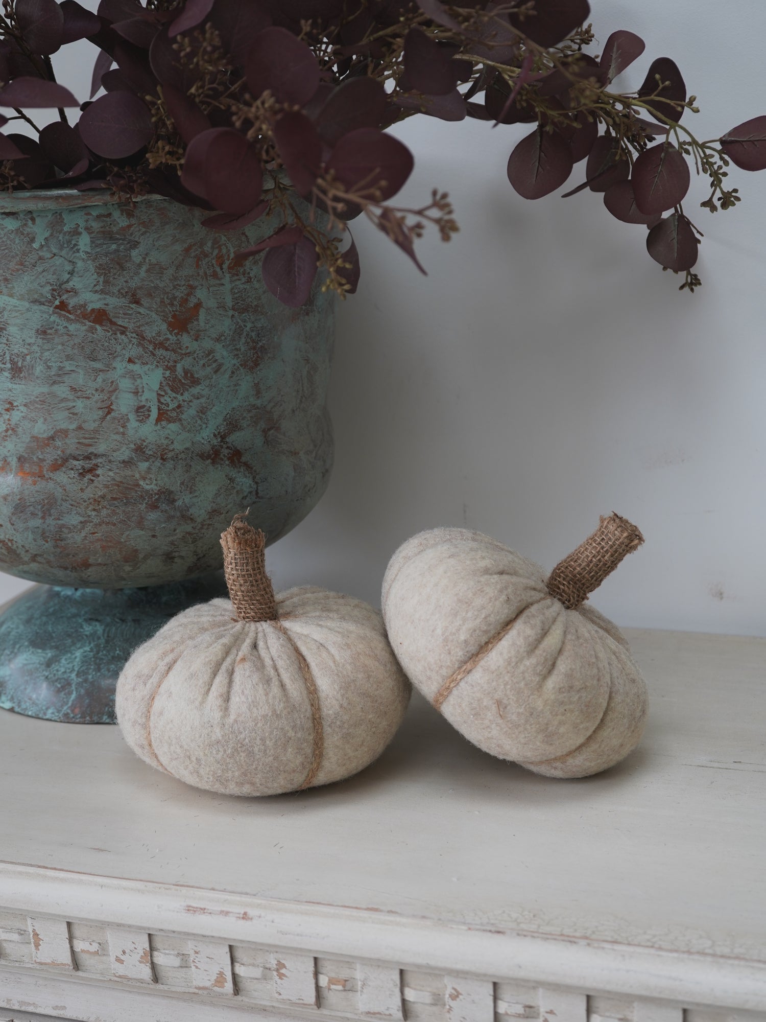 Two beige pumpkins on a white surface with a decorative urn and purple foliage in the background.