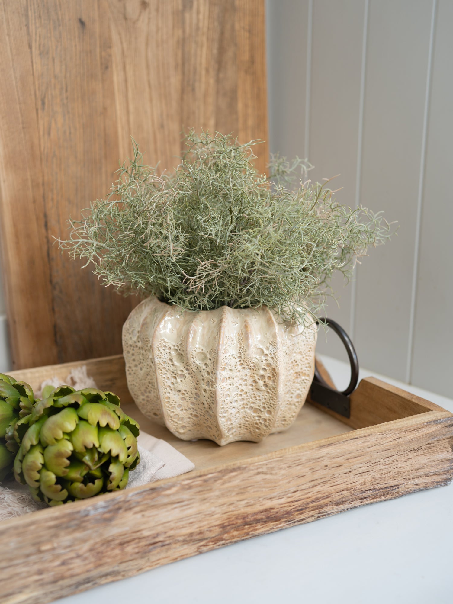 Decorative beige honeycomb effect plant pot with greenery on a wooden tray against a wooden wall.