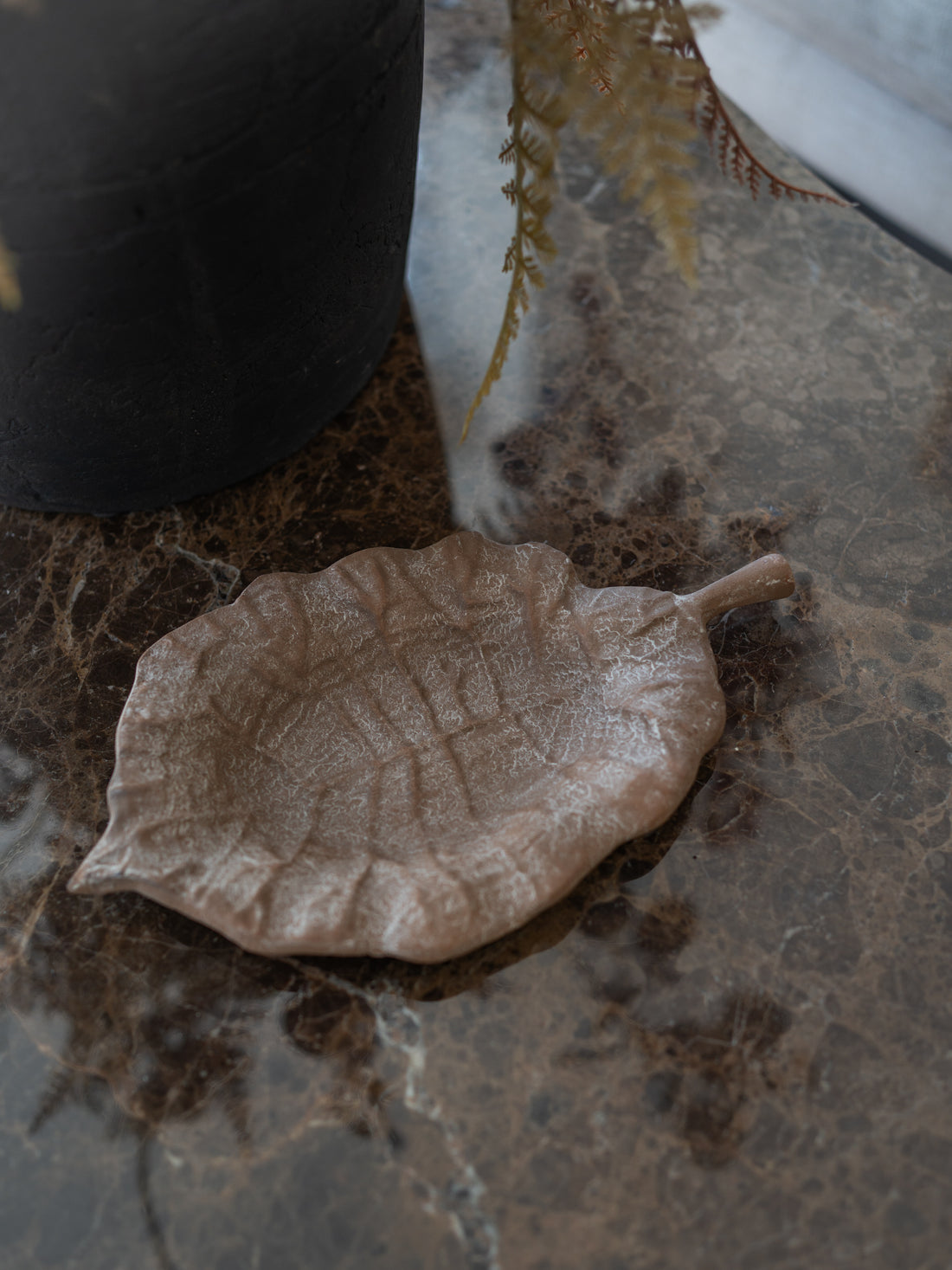 A leaf-shaped ceramic dish in a matte taupe-brown glaze, styled on a dark brown marble surface next to a black planter.