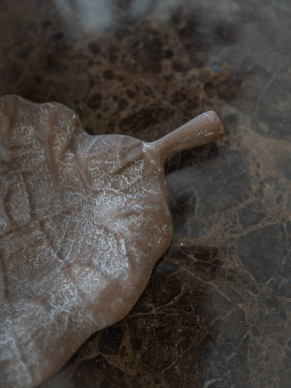 A leaf-shaped metal tray with a textured, matte finish, placed diagonally on a dark brown marble surface.