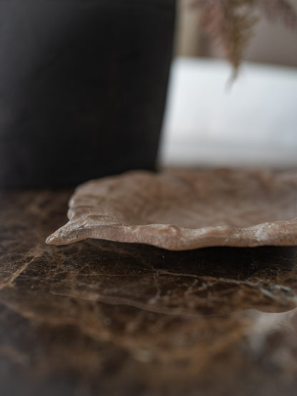A shallow, leaf shaped metal dish in a textured beige and brown glaze, resting on a dark brown marble tabletop with a blurred dark background.