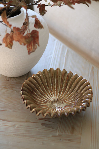 Ceramic ruffle-shaped bowl on a wooden surface with a white vase and dried leaves in the background.