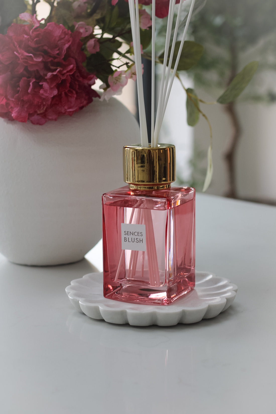 A blush pink reed diffuser with clear liquid and several reeds inserted into it, placed on a white surface.