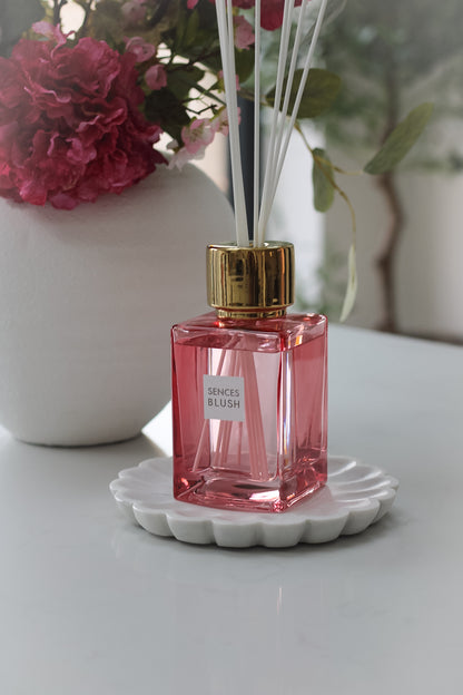 A blush pink reed diffuser with clear liquid and several reeds inserted into it, placed on a white surface.