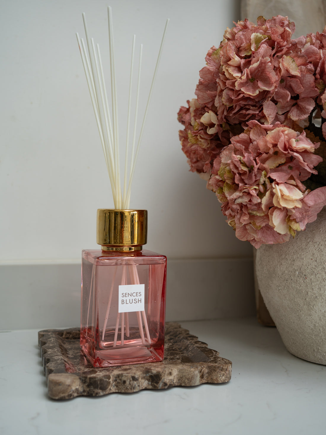 Pink diffuser with gold cap on a brown scalloped marble tray next to pink hydrangeas flowers.