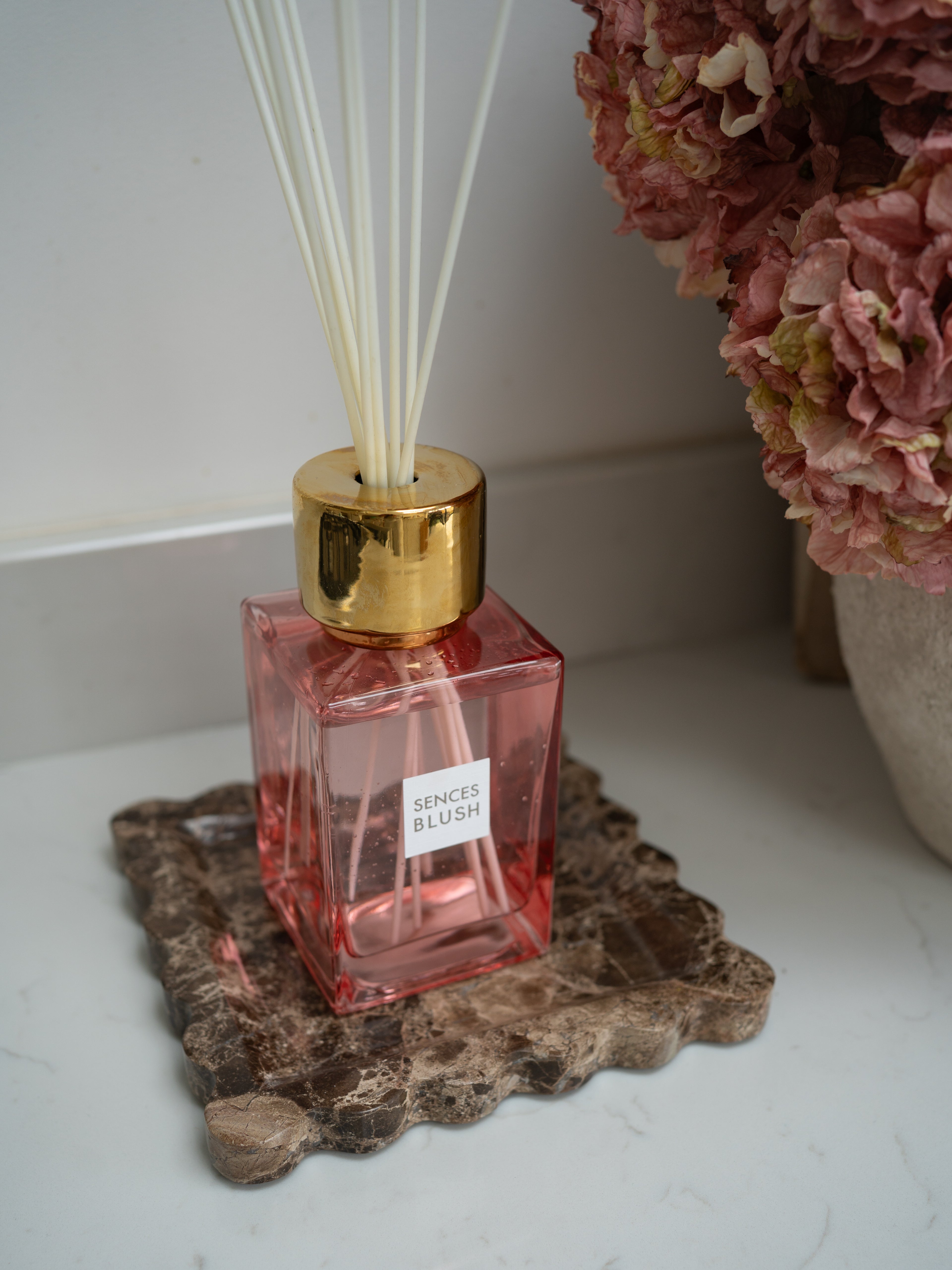 Pink diffuser bottle with gold cap on a brown marble scalloped tray, with pink flowers in the background.