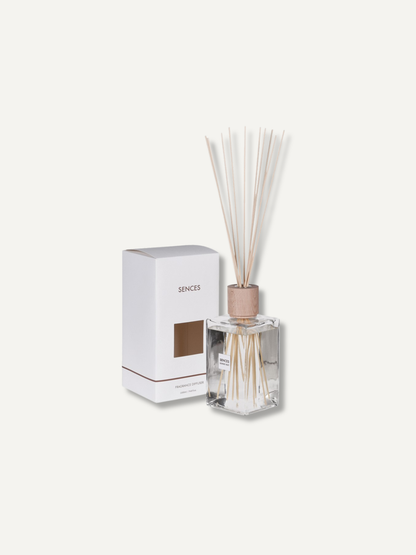 A reed diffuser bottle with light natural reeds and a metallic rose-gold cap, shown next to its white packaging box with the brand name &