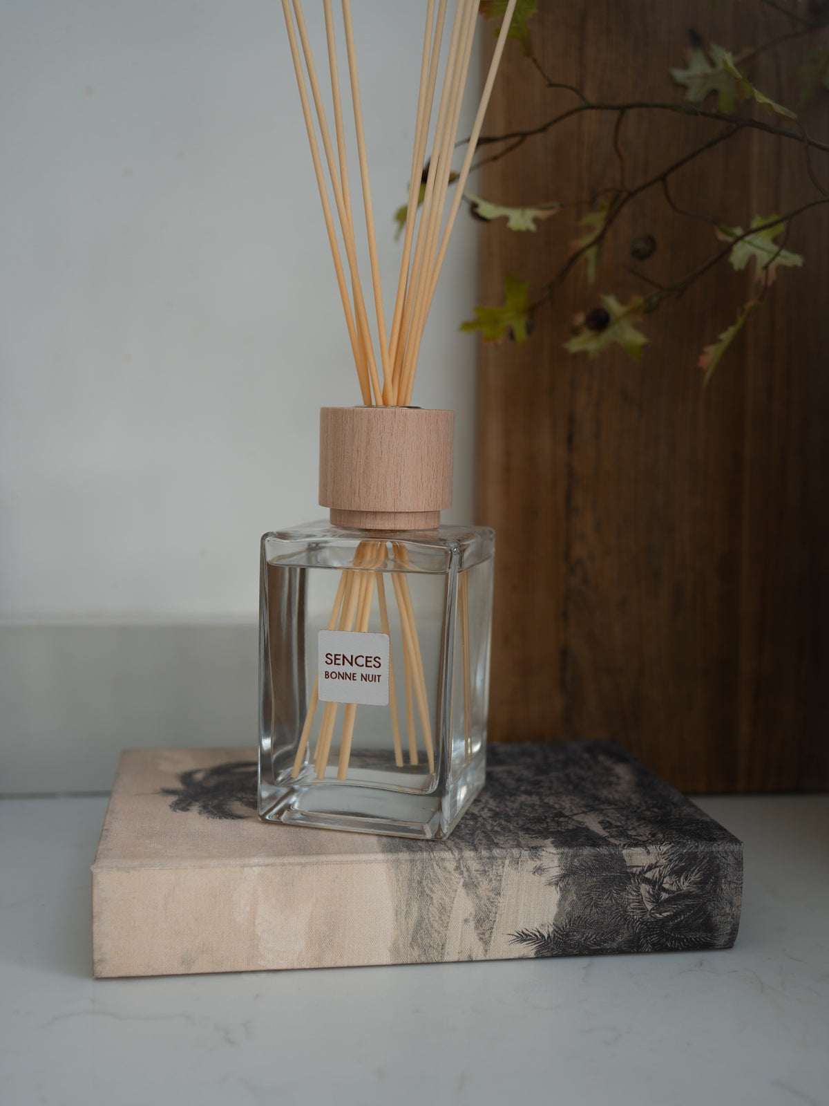 A clear glass reed diffuser bottle with a light wood cap and natural reeds, labeled &