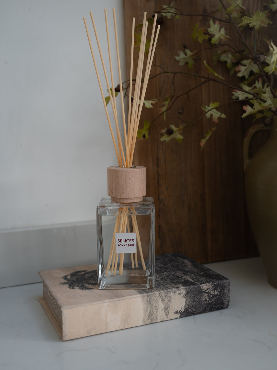 A reed diffuser bottle with natural wood reeds and a matching cap, placed on a decorative book against a white wall and dark wood panel background.