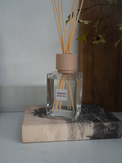 A clear glass reed diffuser bottle with a light wood cap and natural reeds, labeled &