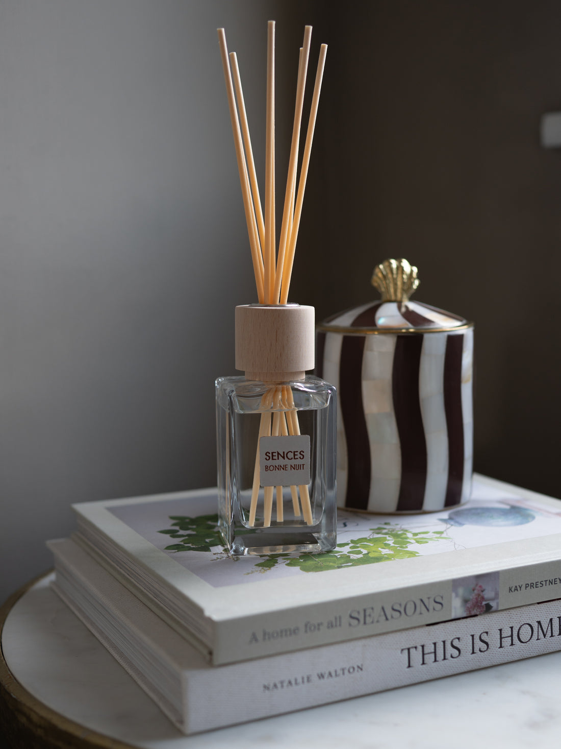 A reed diffuser bottle with light wood reeds and a wooden cap sits on top of two stacked books, with a black-and-white striped ceramic jar behind it against a dark gray wall.