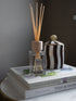A reed diffuser bottle with light wood reeds and a wooden cap sits on top of two stacked books, with a black-and-white striped ceramic jar behind it against a dark gray wall.