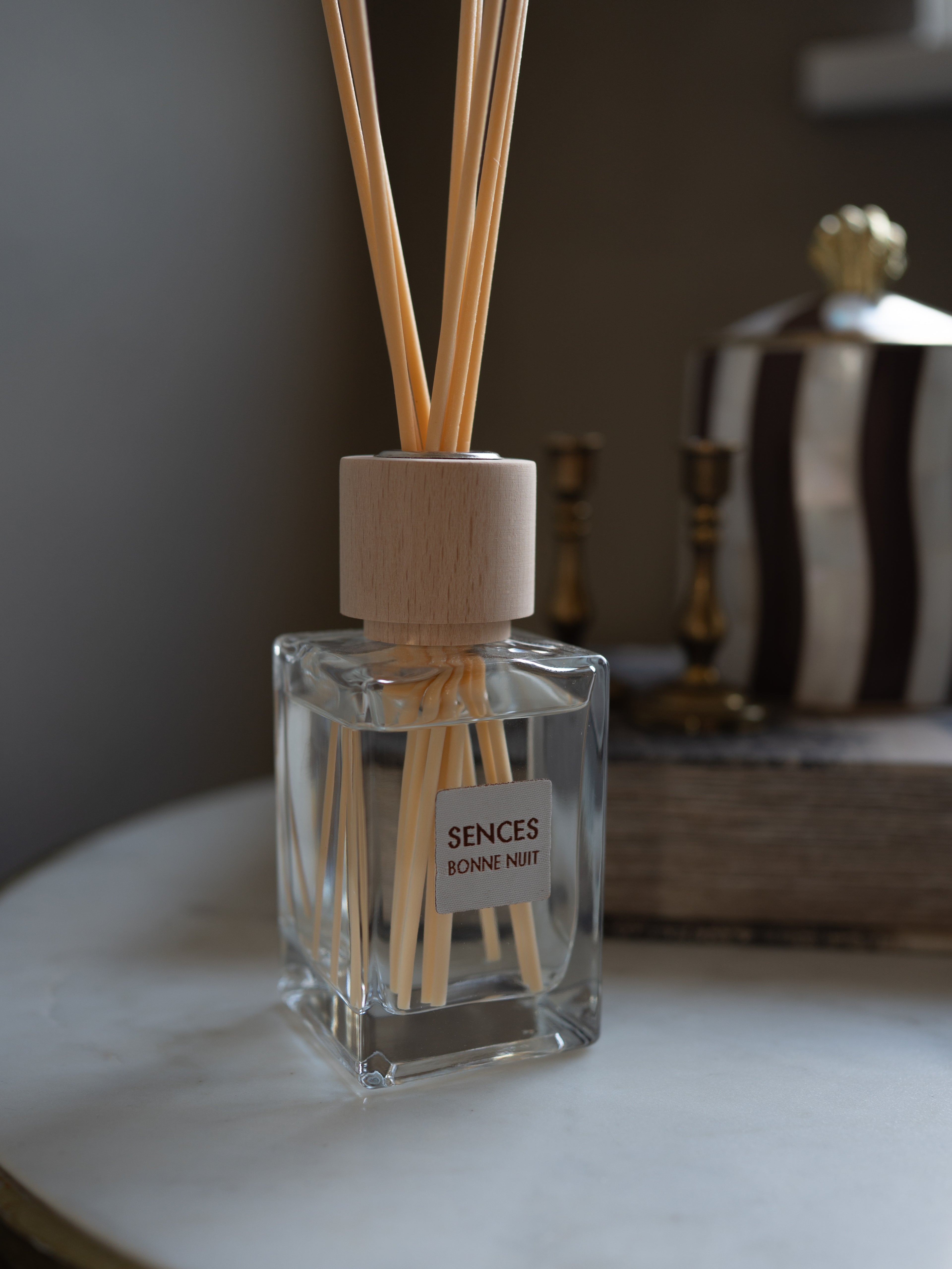 A clear glass reed diffuser bottle with a light wood cap and natural reeds, labeled &